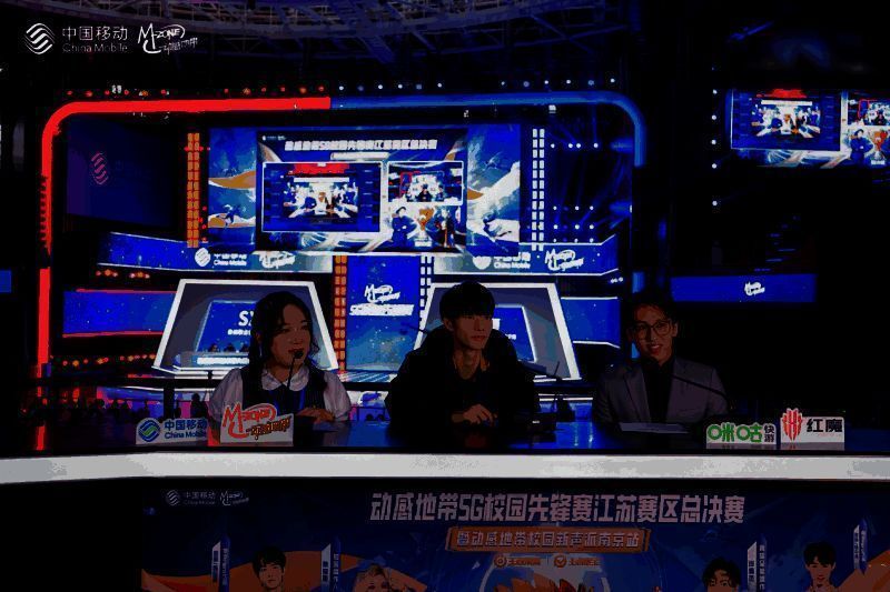 傲寒与景晚在解说台前激情解说,身旁是大屏幕上的比赛画面,现场观众热烈鼓掌